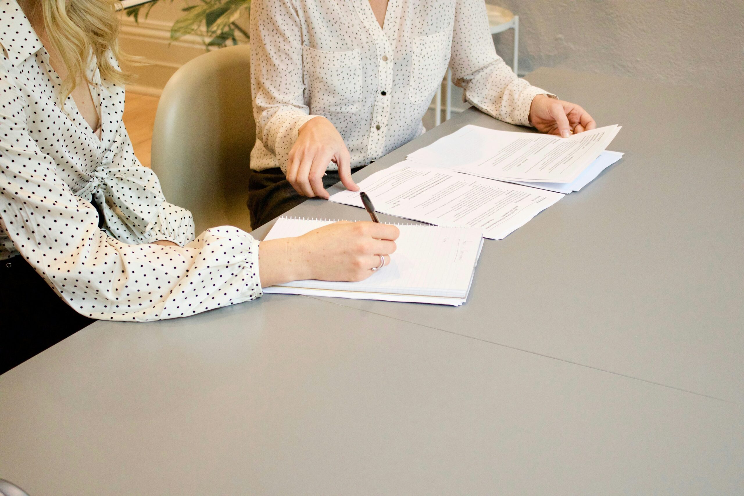 Two Women Working Together To Get Work Delegated via Paperwork sitting at a desk with a pen in their hands.